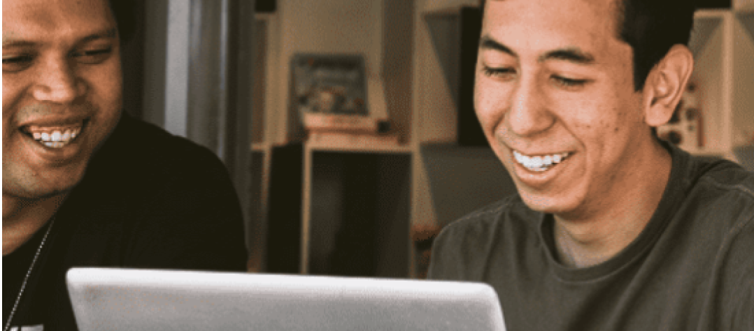 Two smiling individuals collaborating while looking at a laptop screen.